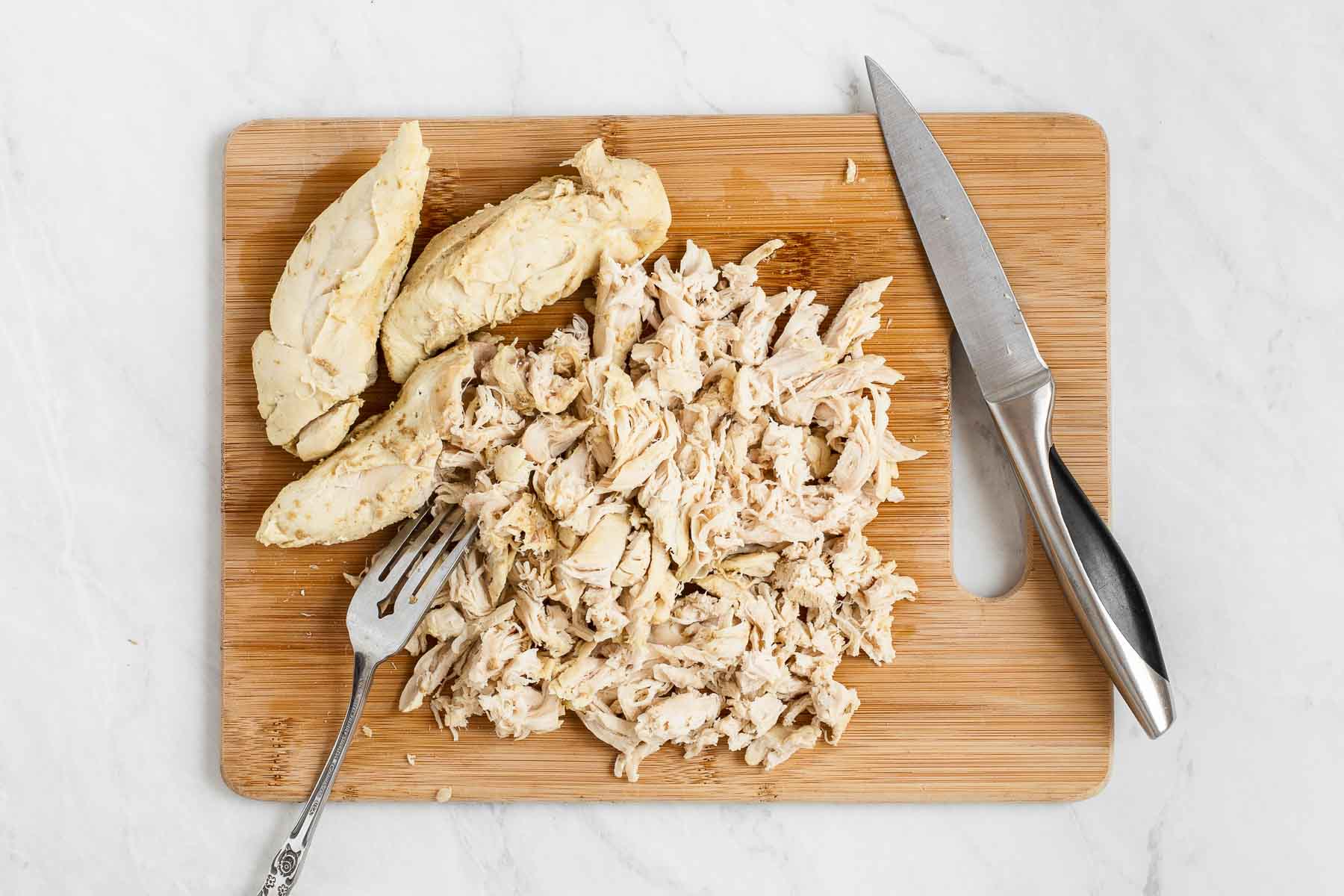 Cooked chicken, being shredded on a wooden cutting board with knife.