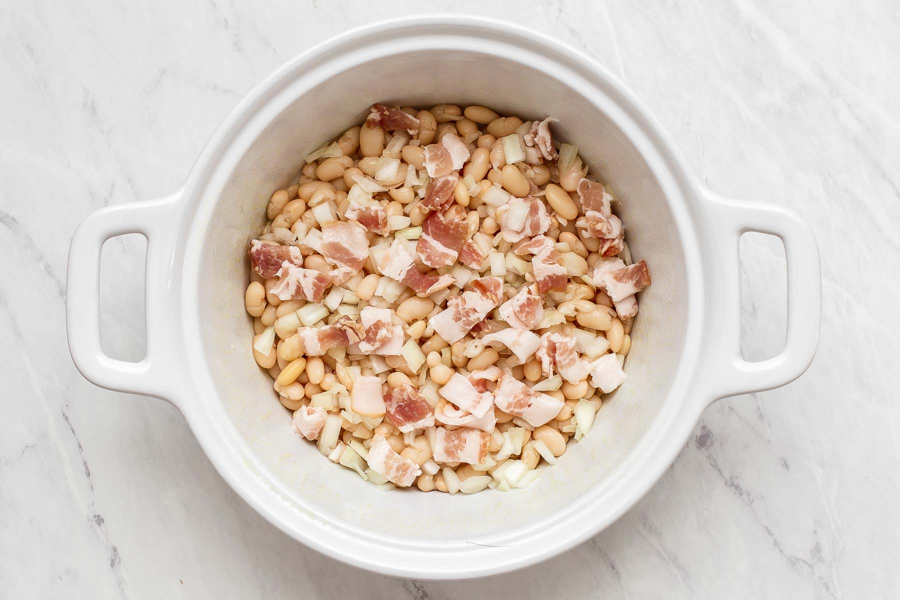 White beans and onions and bacon layered together in white casserole dish.