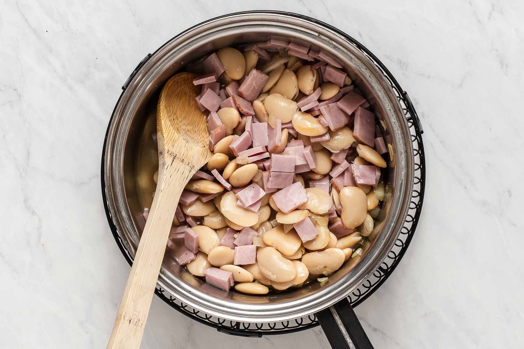 Ham and beans in stainless steel pan with wooden spoon.