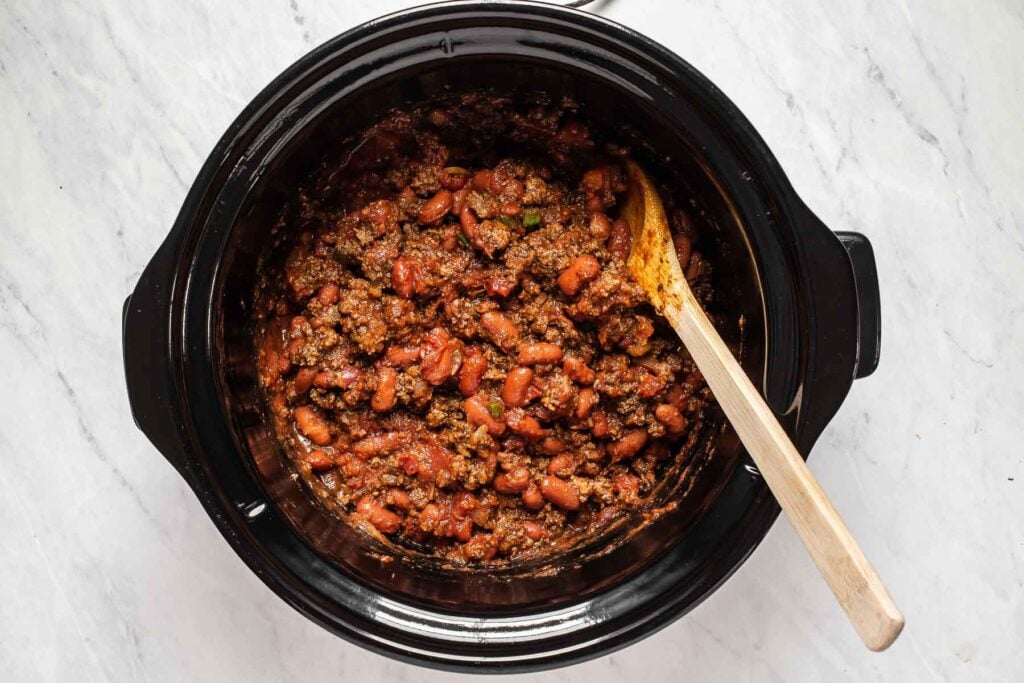 Ground beef, tomatoes and beans in a black slow cooker with wooden spoon.