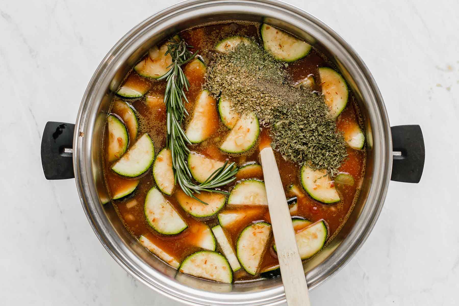 Dried herbs and rosemary sprig in soup pot with zucchini and tomato broth.