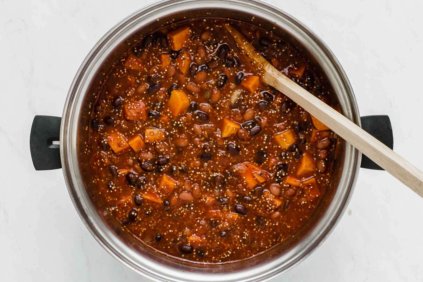 Sweet potato and bean chili cooking in pot with wooden spoon.
