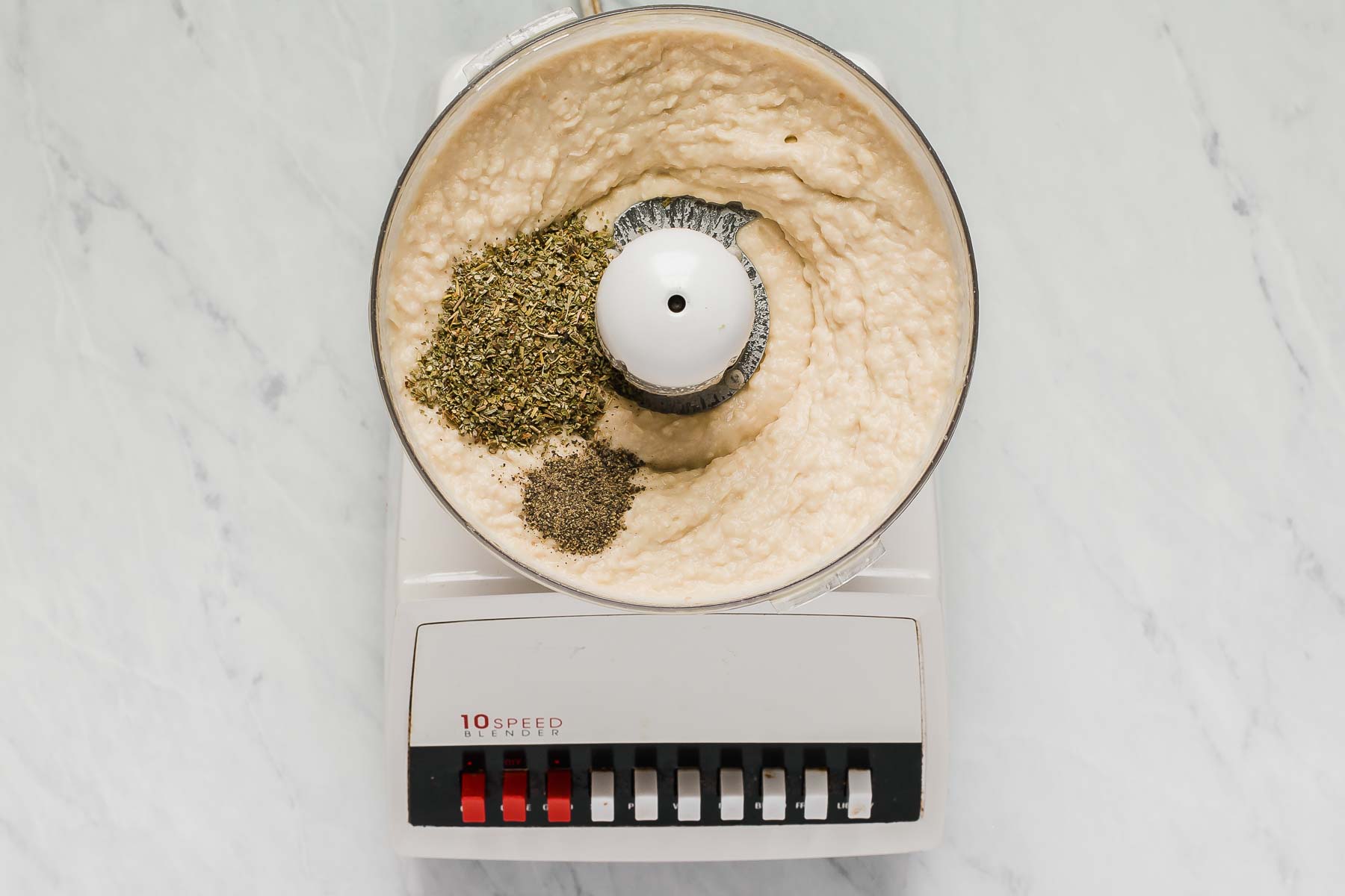 White bean dip in food processor with herbs on top.