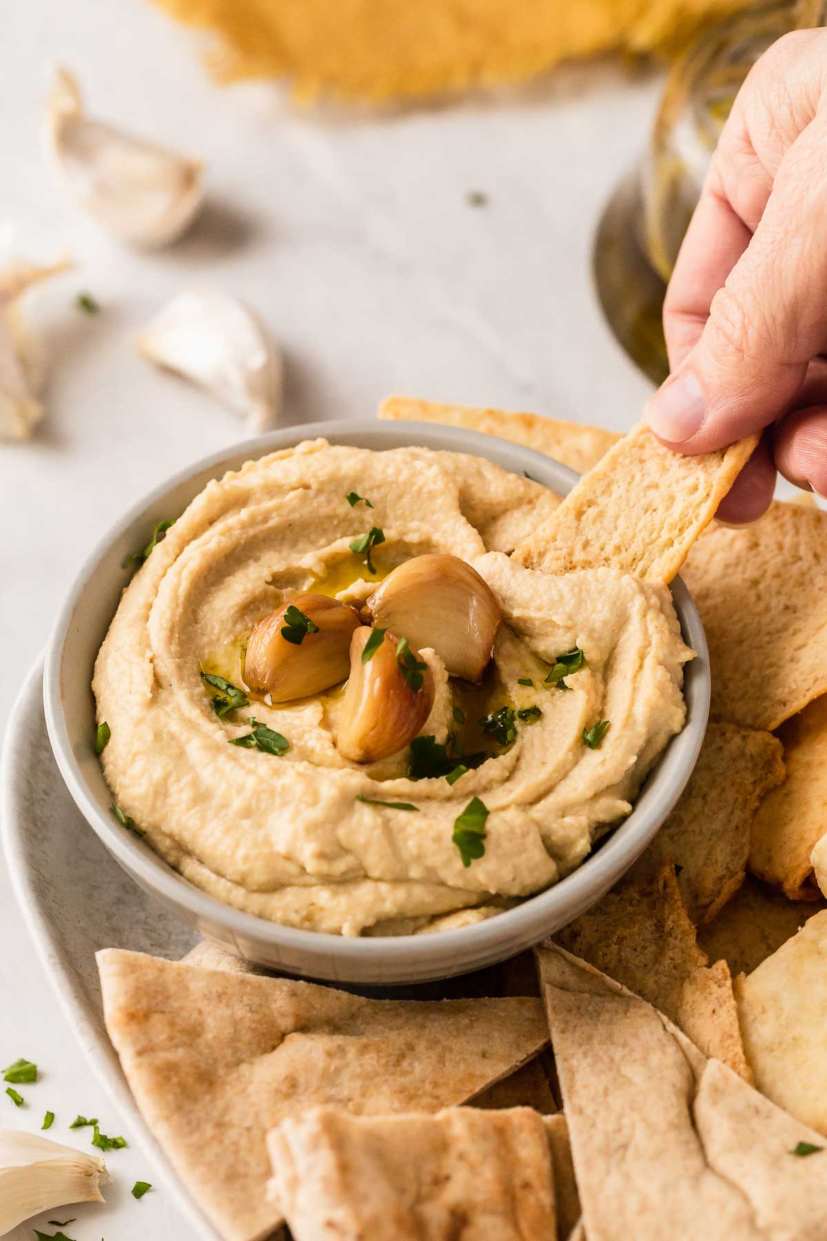 Hand dipping pita chip into hummus with garlic cloves on top.