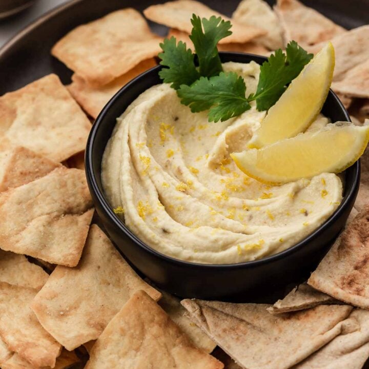 Bowl of butter bean hummus surrounded by pita chips and lemon wedges.