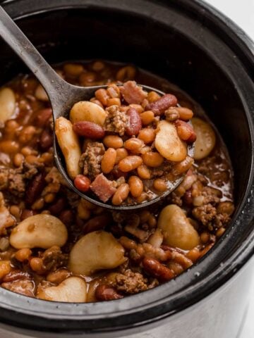 Black crockpot full of mixed beans and ground beef with spoon removing a scoop.