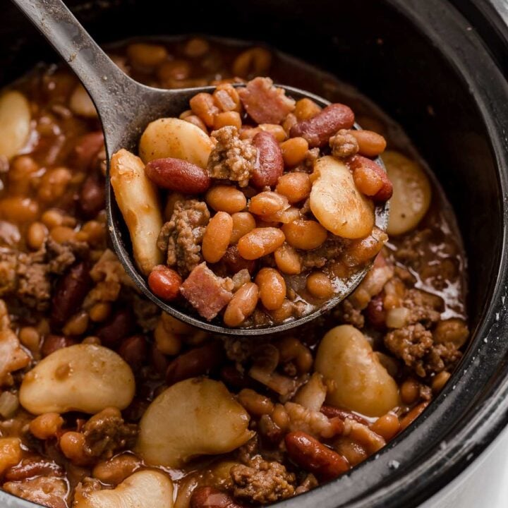 Black crockpot full of mixed beans and ground beef with spoon removing a scoop.