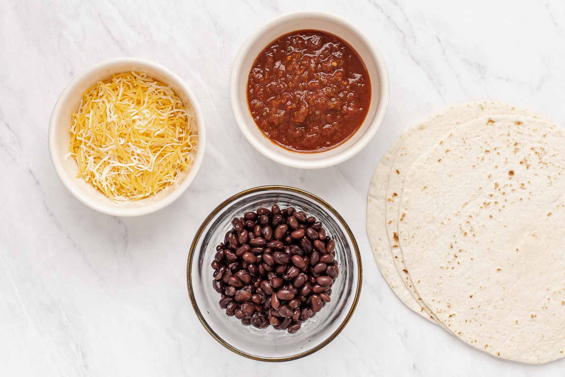 Bowl of cheese, salsa, black beans and tortilla on white table.