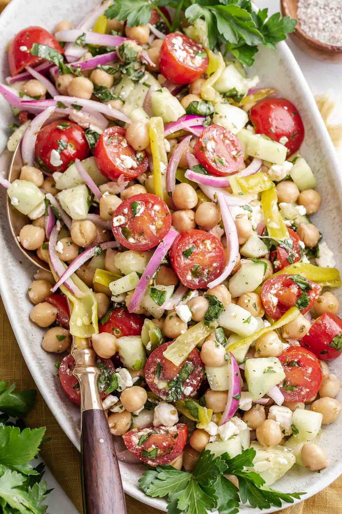 Tossed Mediterranean chickpea salad on oval serving platter with tongs.