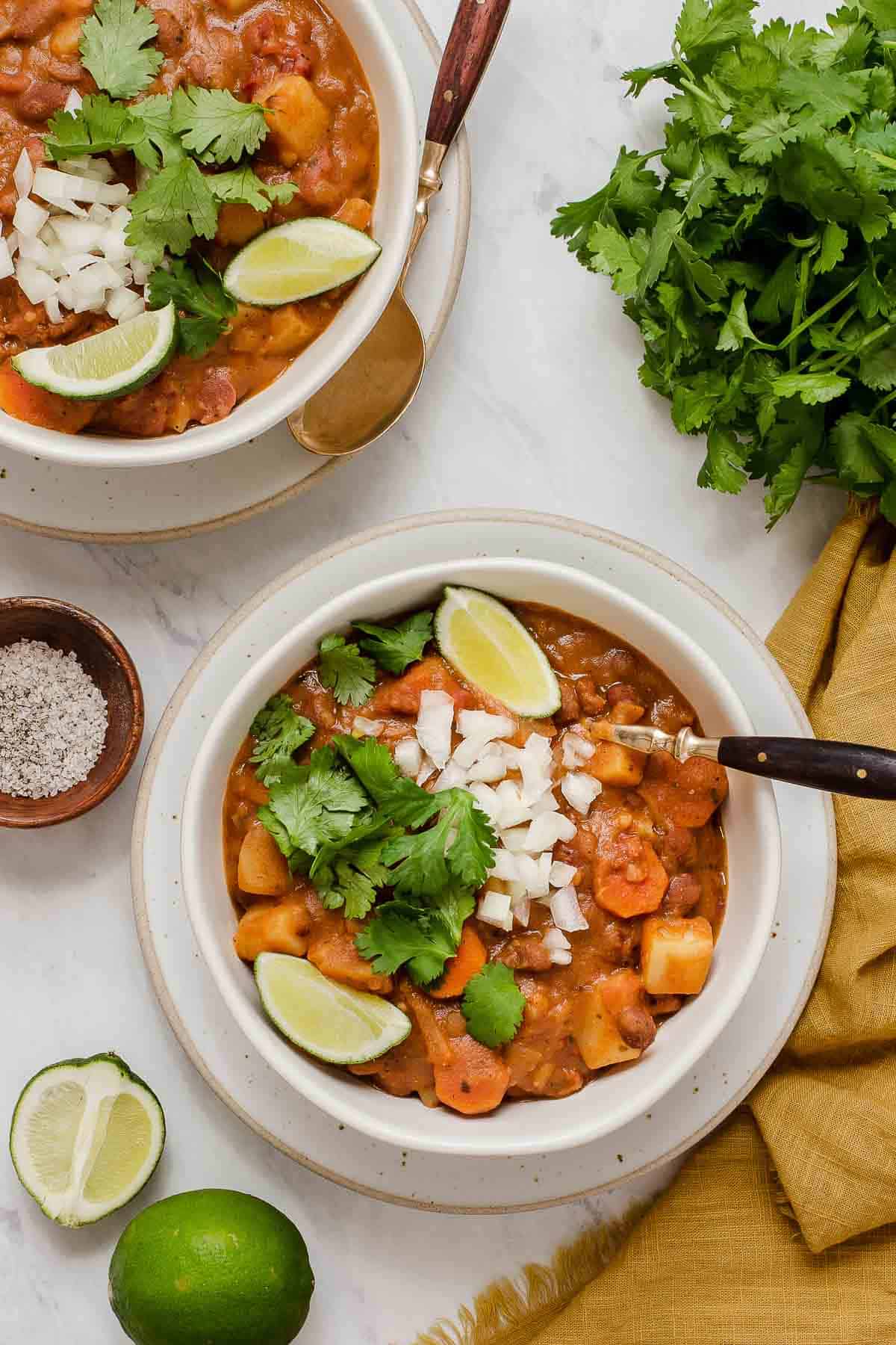 Vertical photo of two bowls of pinto bean soup garnished with cilantro, lime and diced onion.