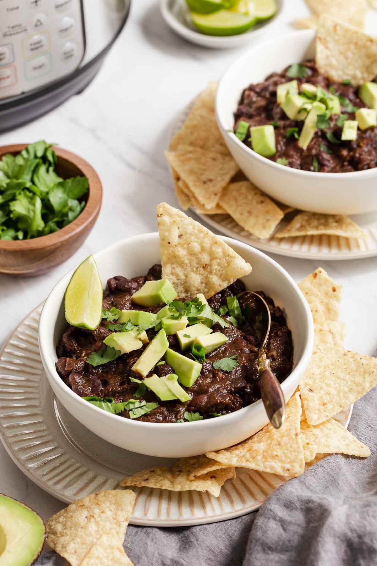 White table with two bowls of black bean chili garnished with chips and avocado chunks.