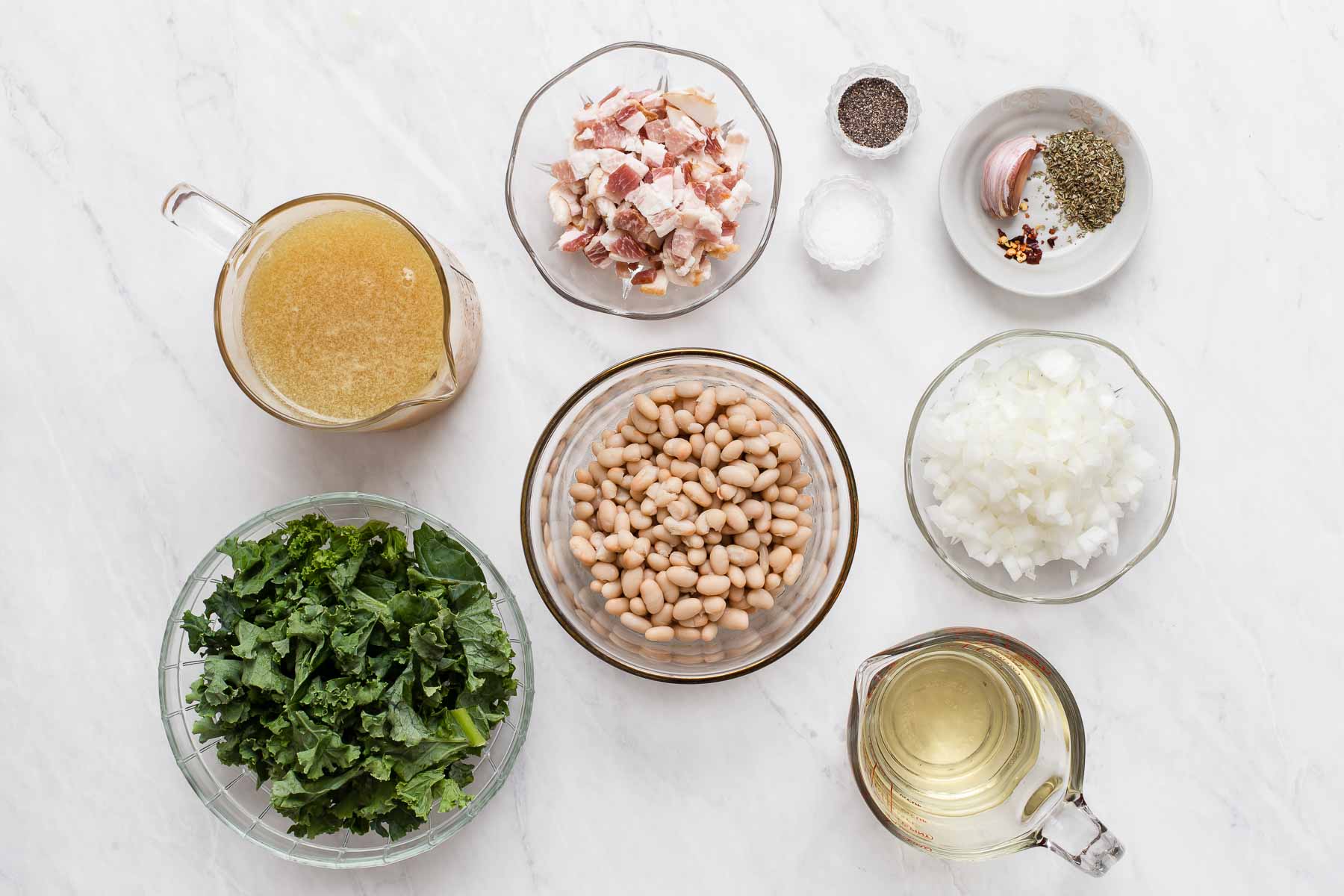 White beans, kale, chicken broth, and diced bacon in small bowls on white counter.