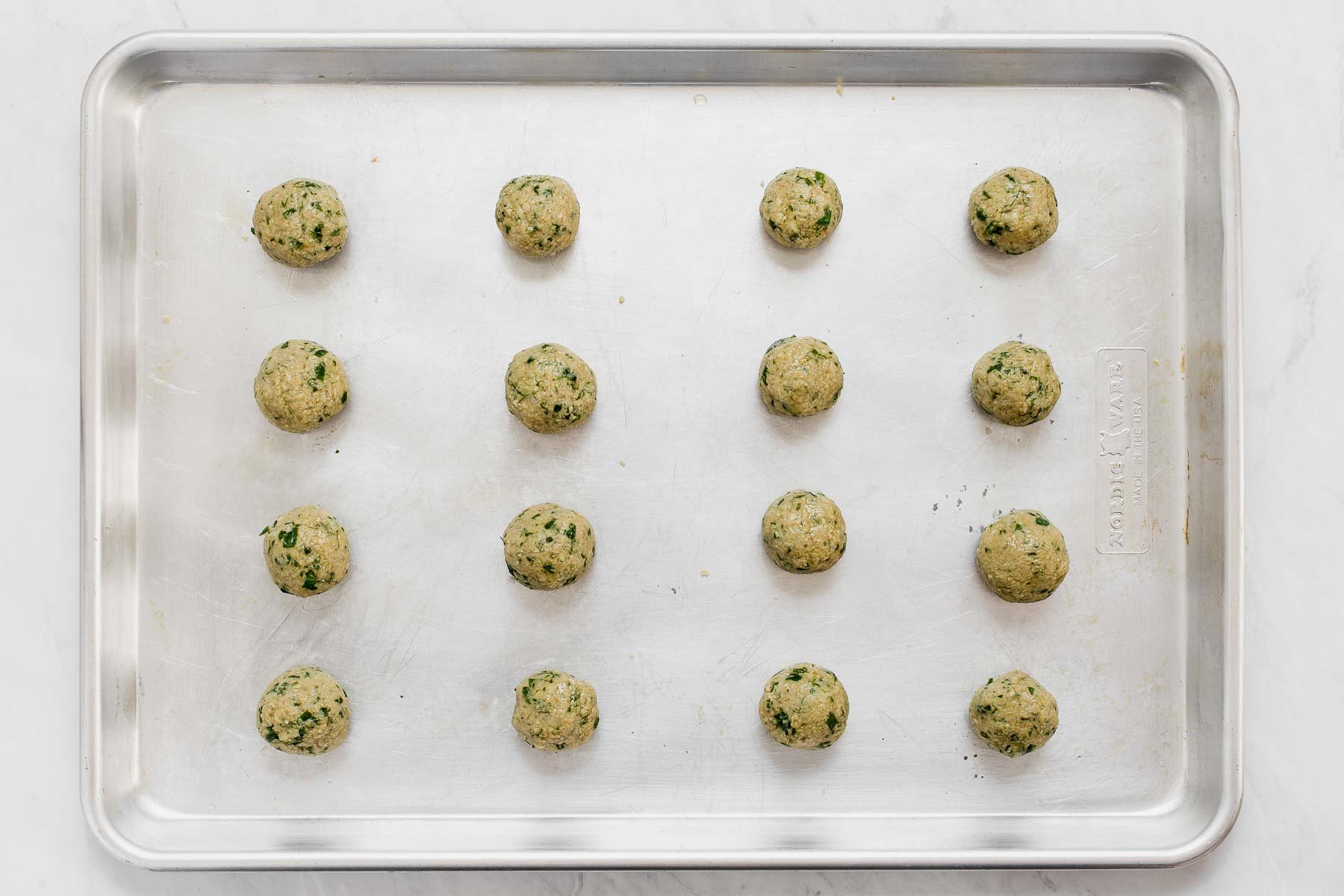 Sixteen brown and green balls of raw falafel batter on a baking sheet lined with parchment paper.