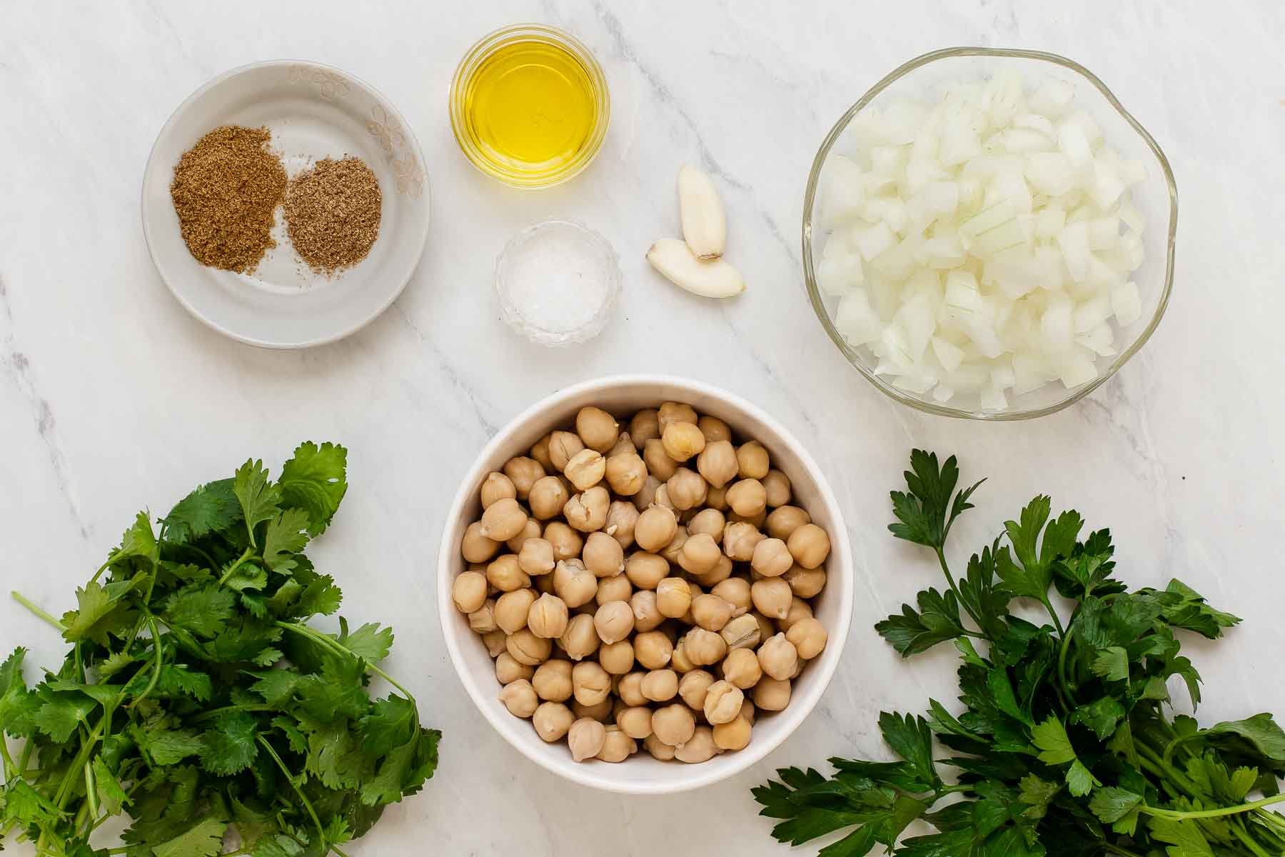 Chickpeas, spices, oil, chopped onion and herbs in small bowls on white counter.