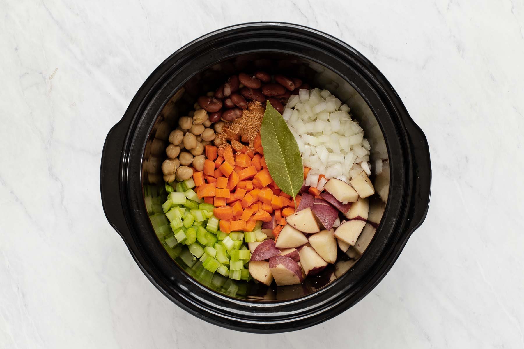 Raw diced carrots, celery, beans and bay leaf in a black slow cooker.