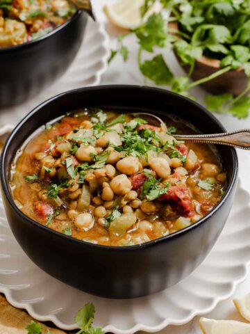Black bowl with Moroccan chickpea stew with lentils, tomatoes and greens on side.