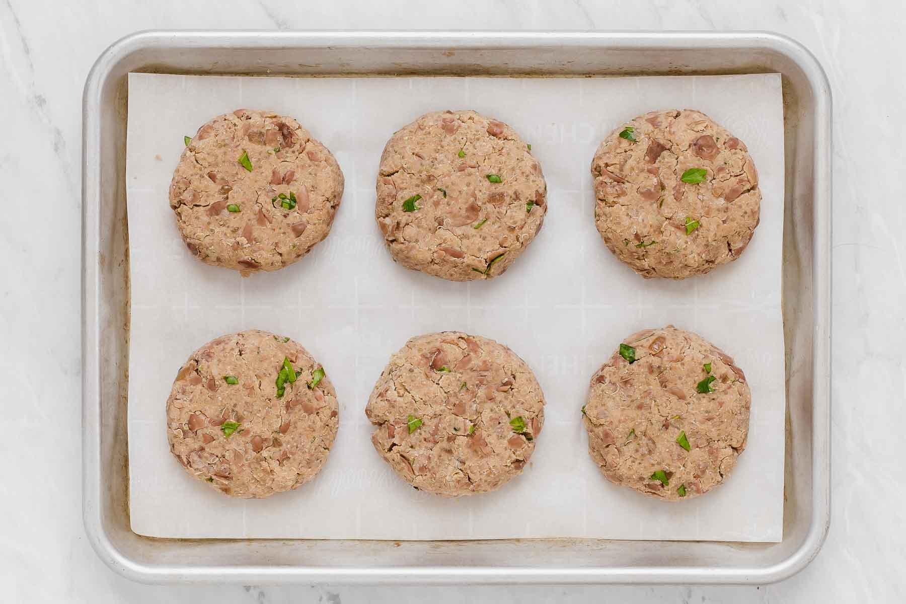 Six pinto bean burgers on a baking sheet before being cooked.