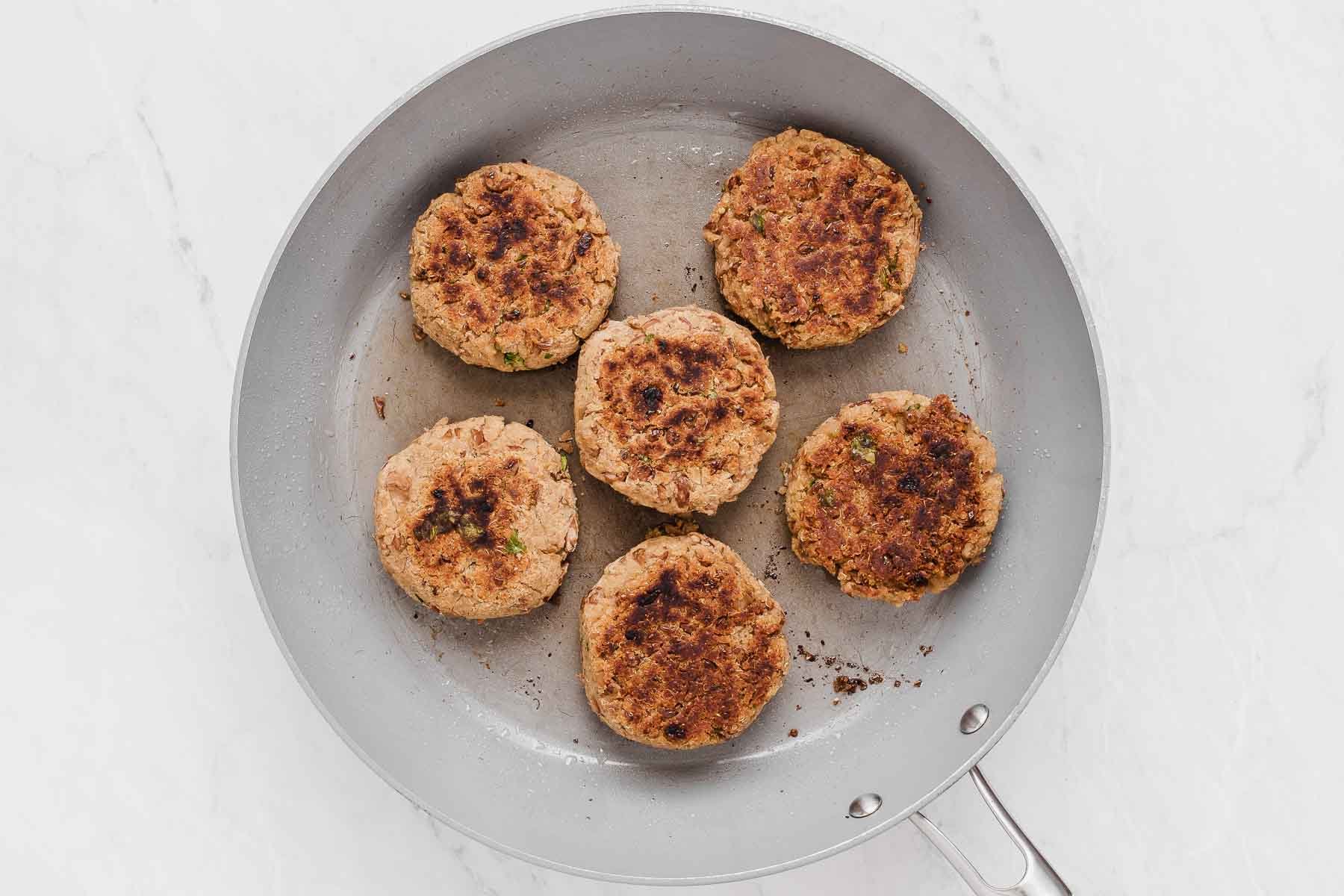 Six pinto bean burgers cooked to golden brown on one side in a grey skillet.