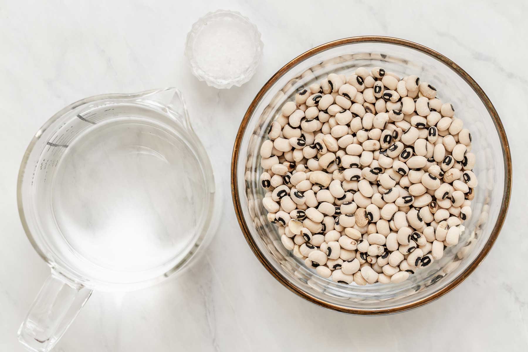 Horizontal image of dried black eyes peas, a cup of water and small dish of salt.