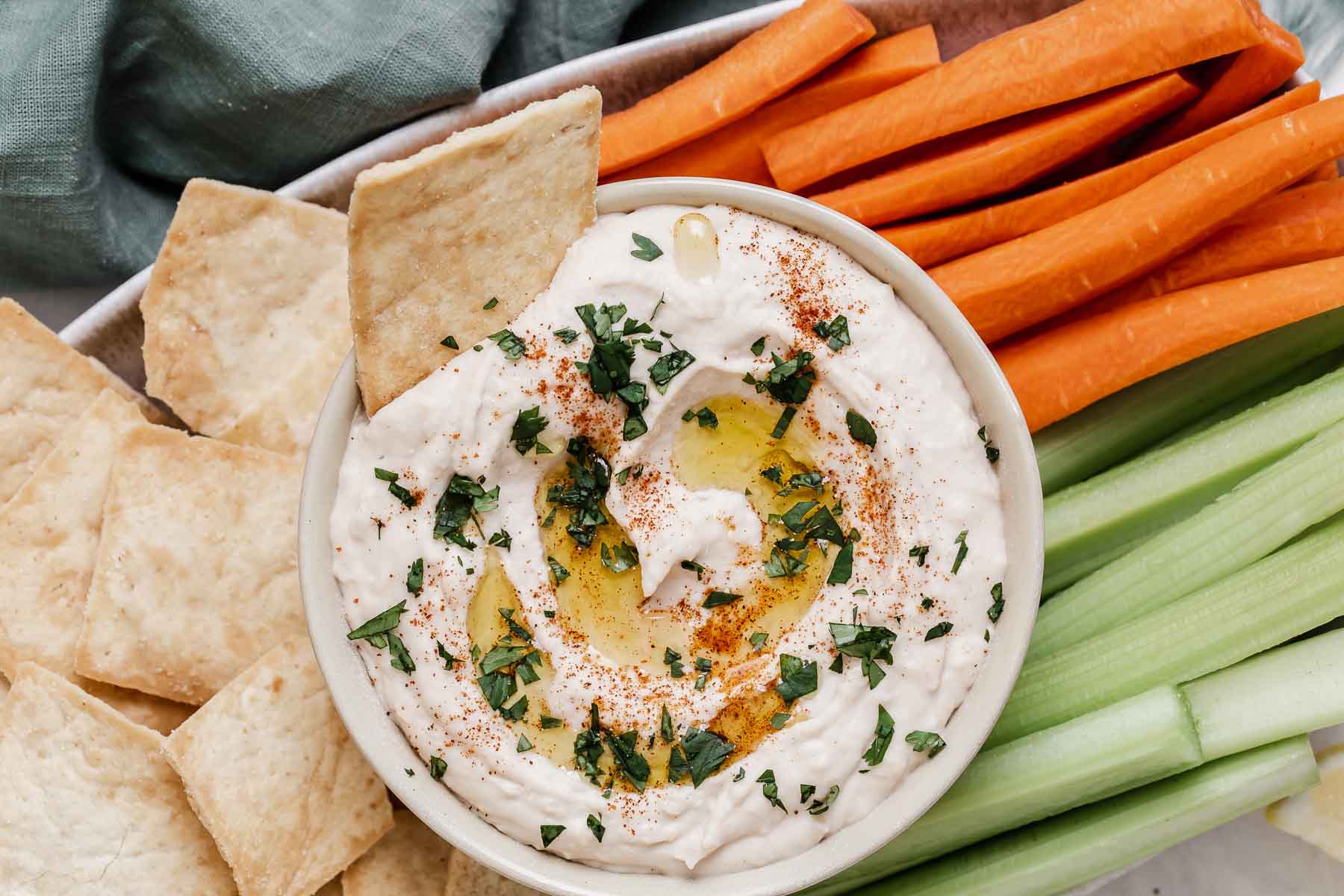 Bowl of white bean hummus topped with olive oil and herbs, surrounded by crudite for dipping.