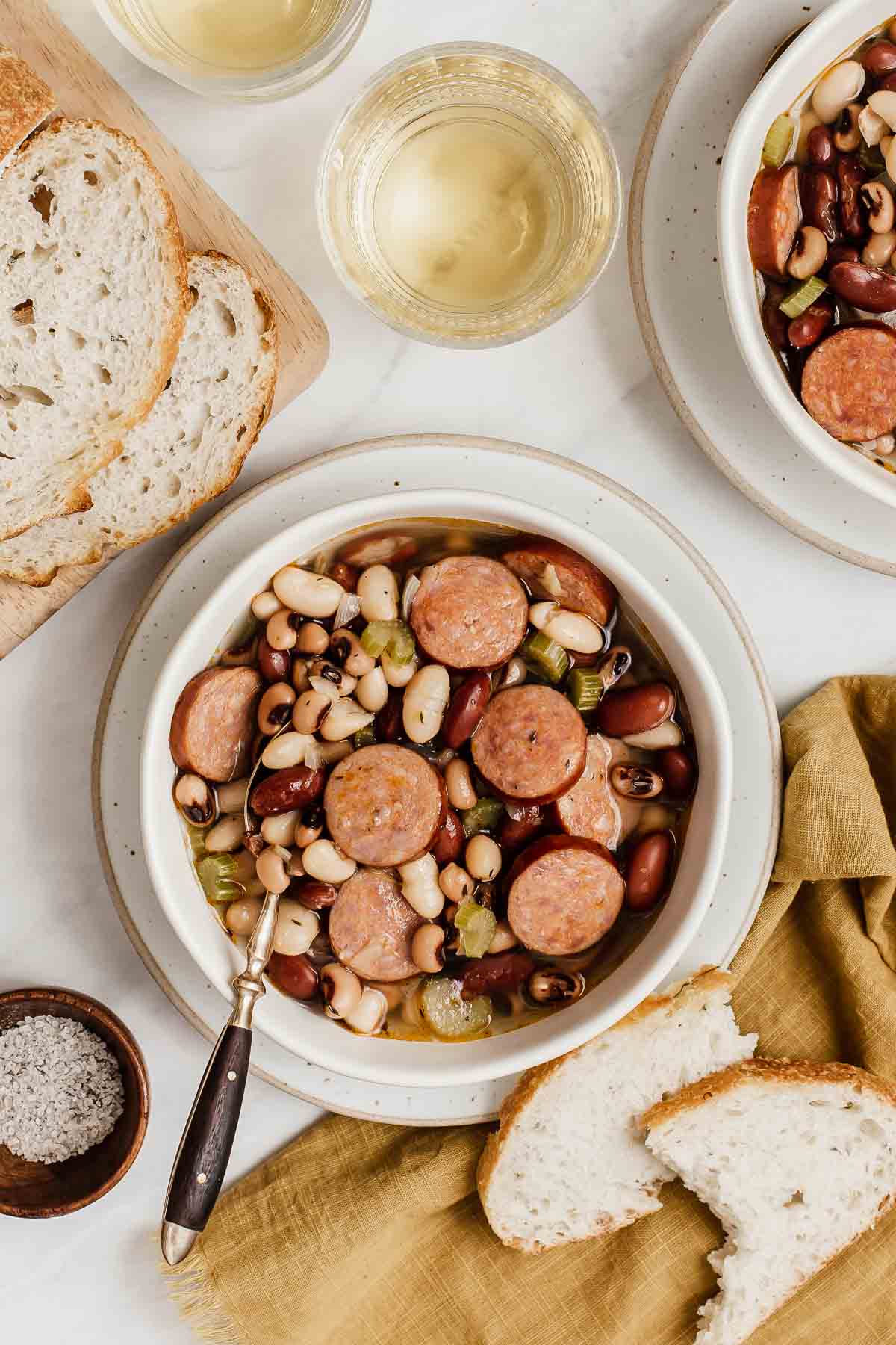 Vertical image of one bowl of 3 bean soup with sausage and glass of wine on side.