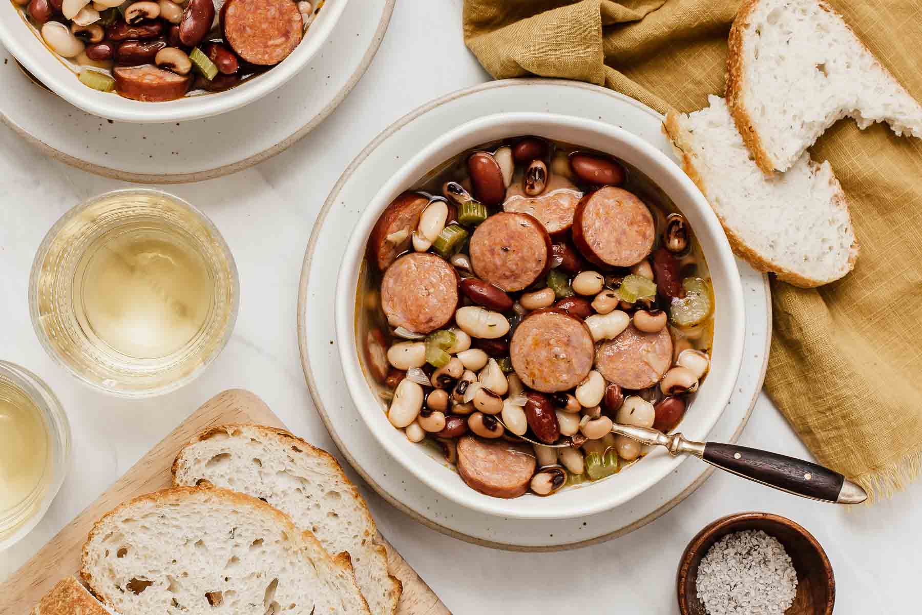 Bowl of 3 bean soup with sliced sausage in white bowl with bread and wine on the side.