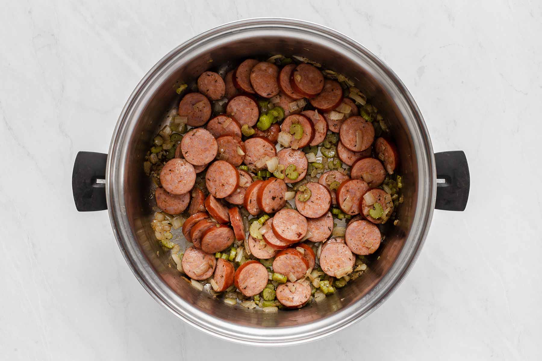 Soup pot with sliced sausage sautéing on top of onions and celery and herbs.