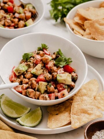 Chickpea salad in white bowl with limes and tortilla chips on side.