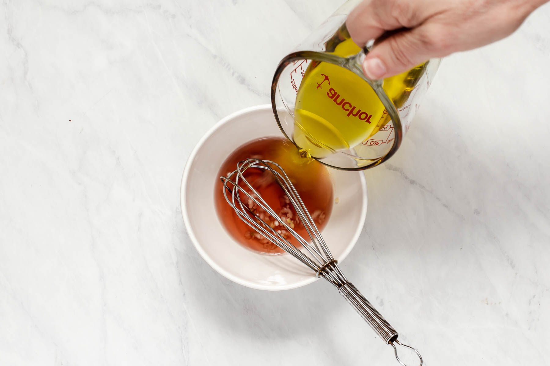 Emulsifying olive oil into a vinaigrette in a small white bowl with whisk.