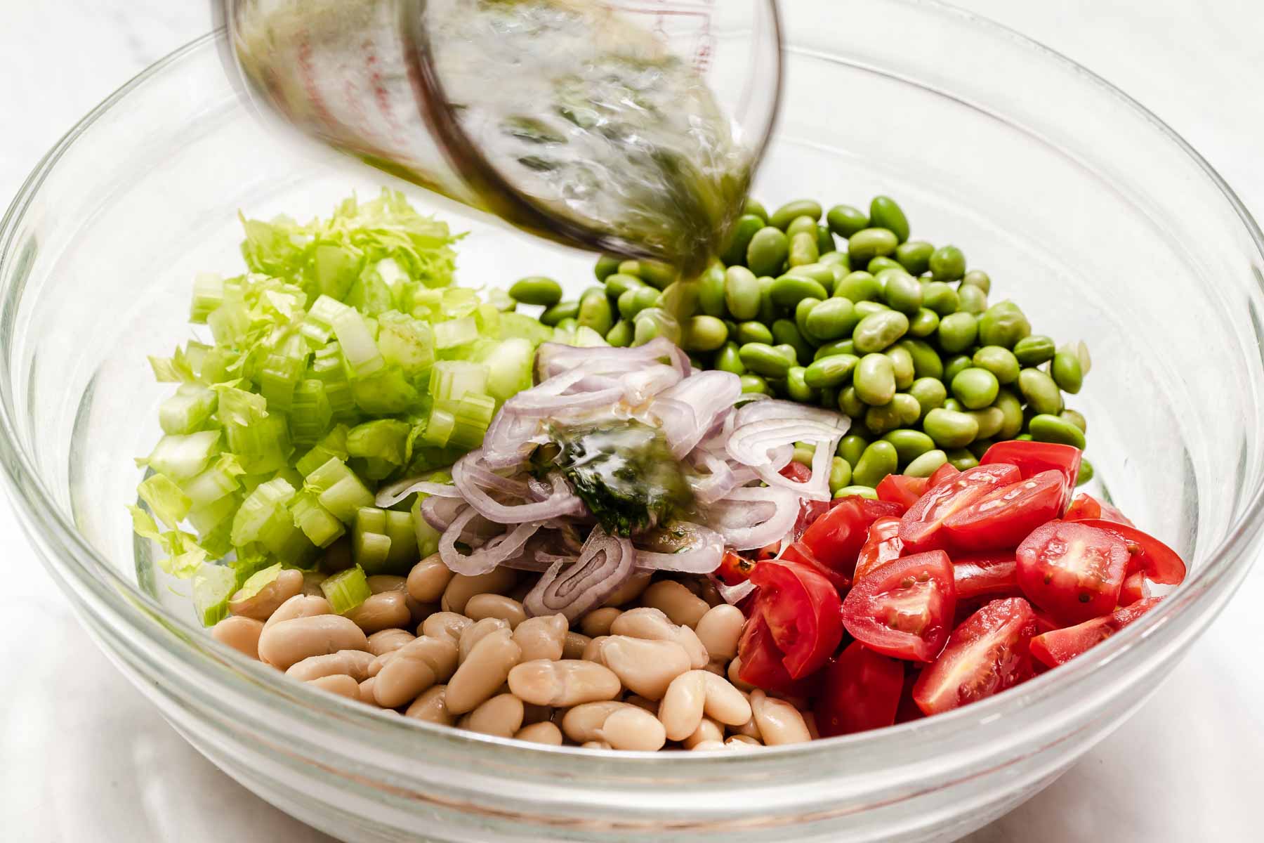 Pouring vinaigrette over edamame white bean salad in clear bowl.