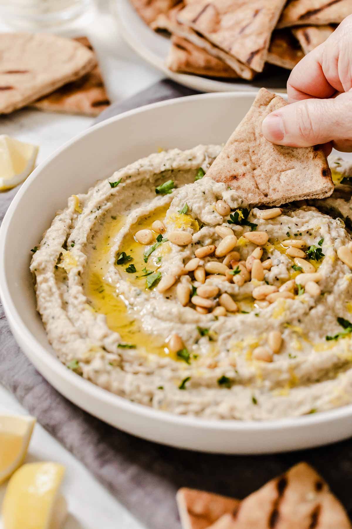 Hand dipping pita triangle into dip with olive oil and pine nuts on top.
