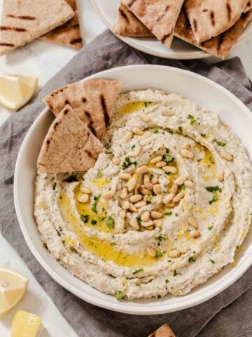 Bowl of white bean eggplant dip with herbs, pine nuts and pita triangles on side.