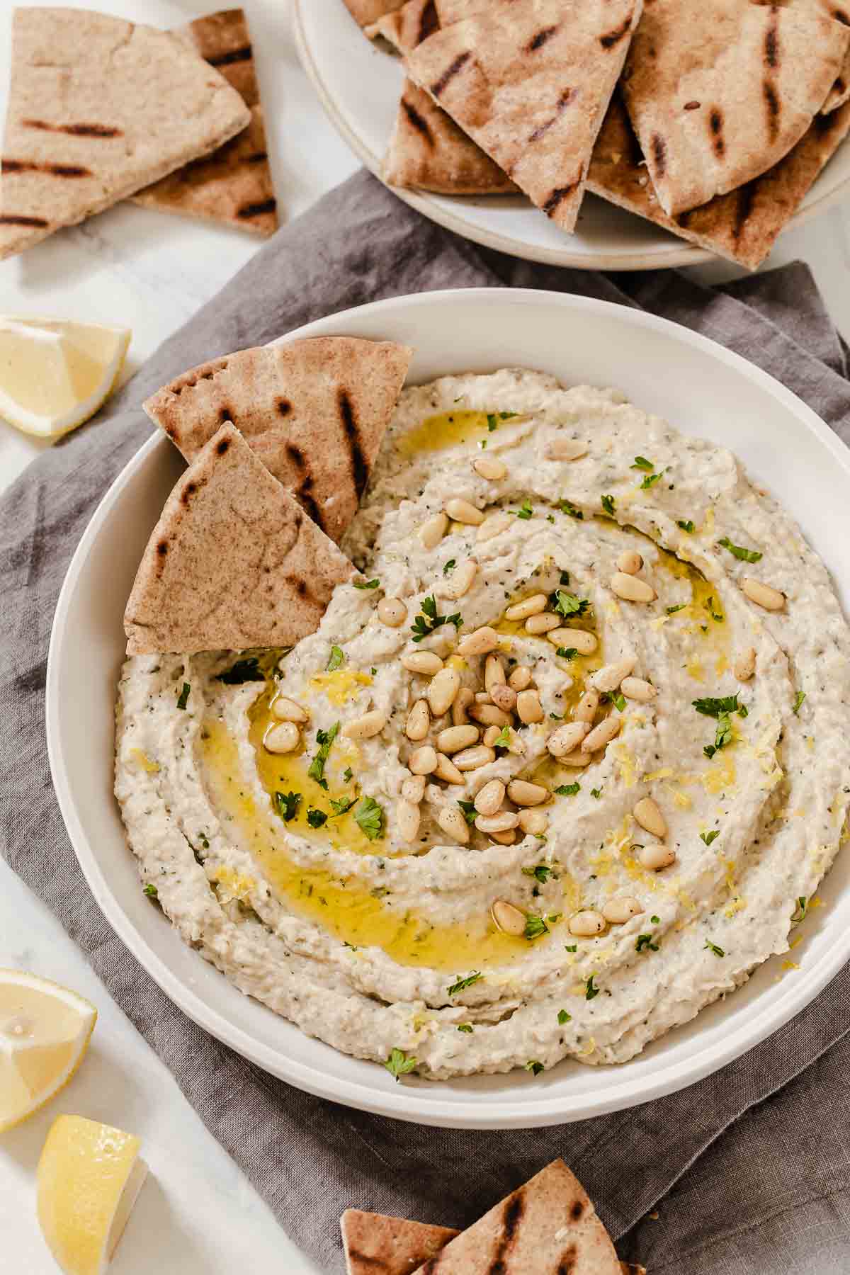Bowl of white bean eggplant dip with herbs, pine nuts and pita triangles on side.