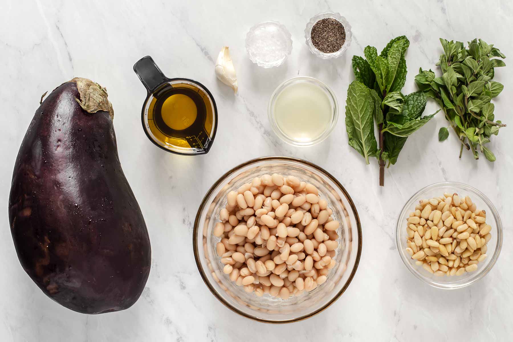 Eggplant, white beans, fresh herbs and pine nuts on marble counter.