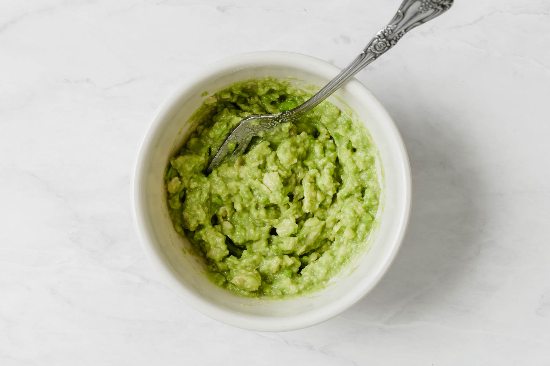 Green mashed avocado in small white bowl with fork on side.