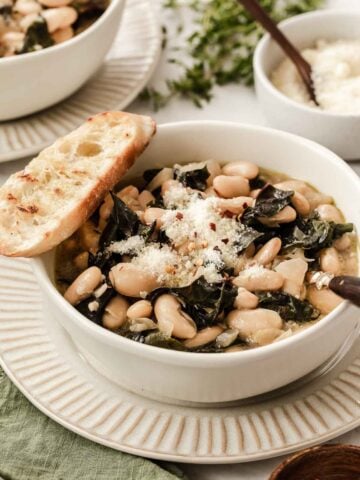 Bowl of white beans and greens with Parmesan cheese and bread slice on top.