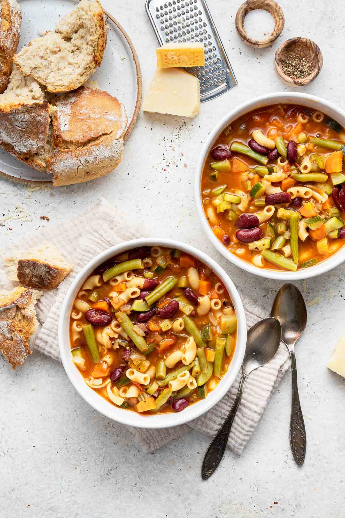 Two white bowls of minestrone soup with crusty bread on the side.