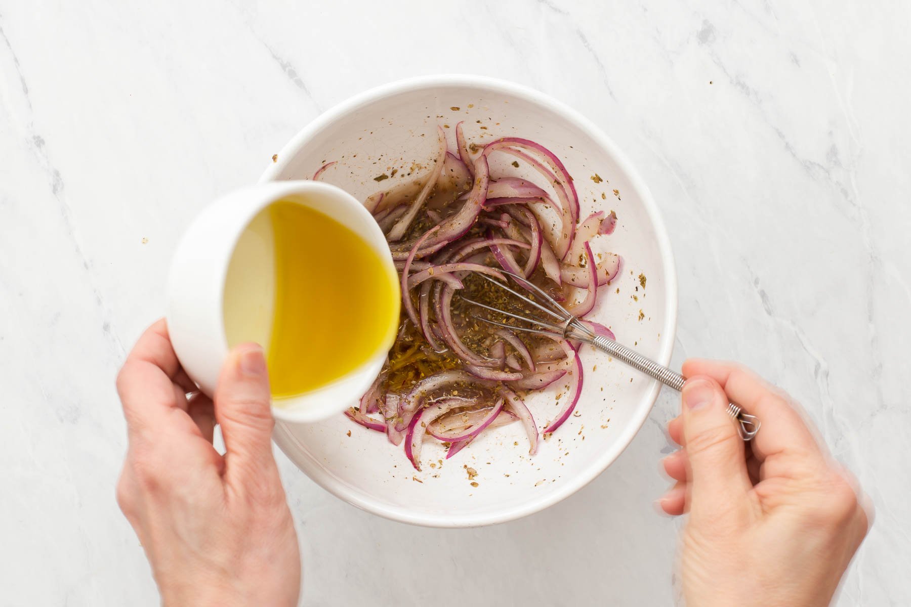 Hand pouring olive oil in bowl with red onions and herbs.