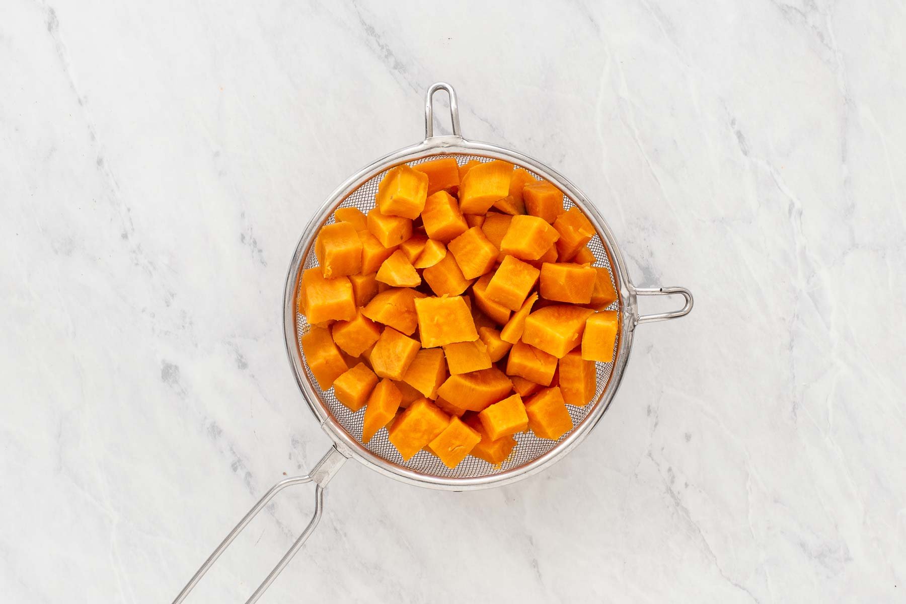 Cooked sweet potato draining in a colander on marble counter top.
