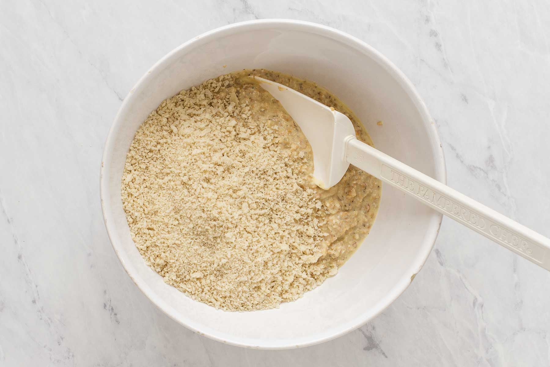 Bowl with bread crumbs and yellow mixture being stirred by white spatula.