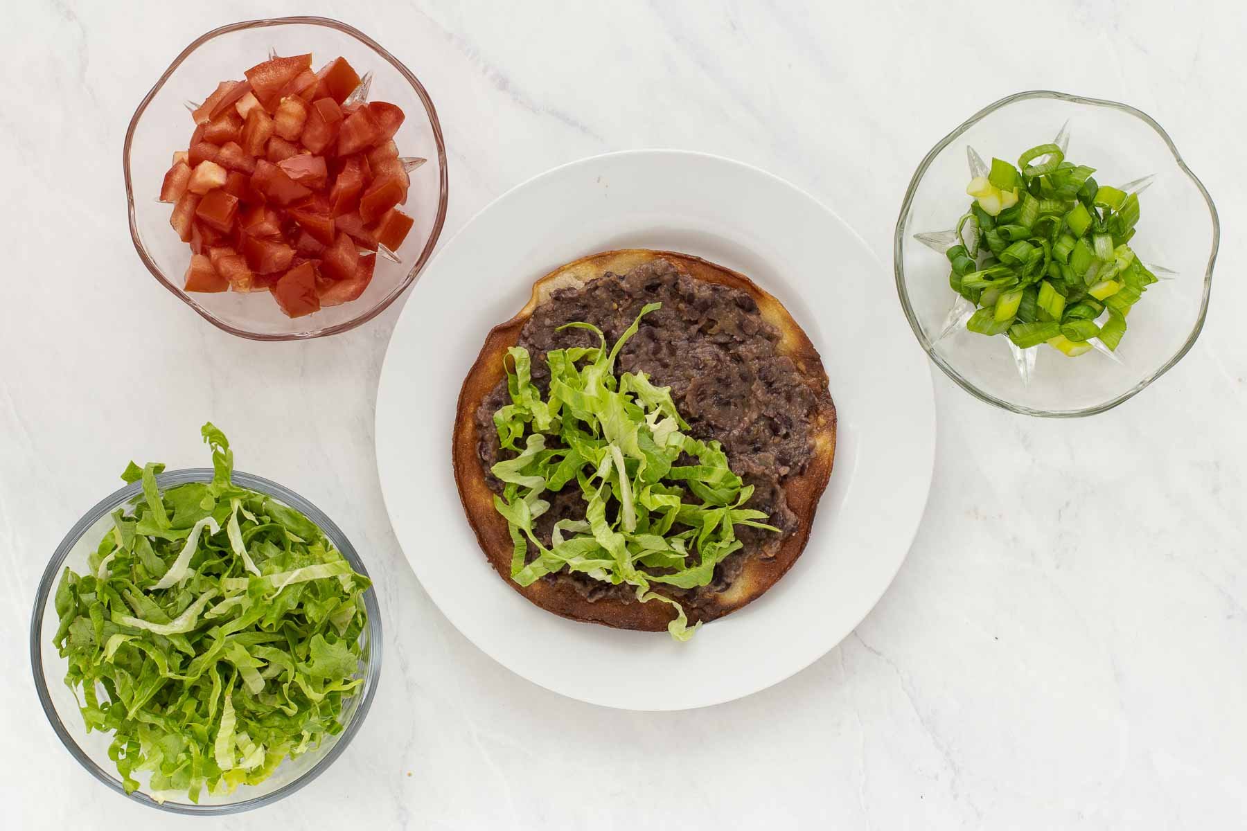 Layering lettuce and chopped tomatoes on a black bean tostada on a white plate.