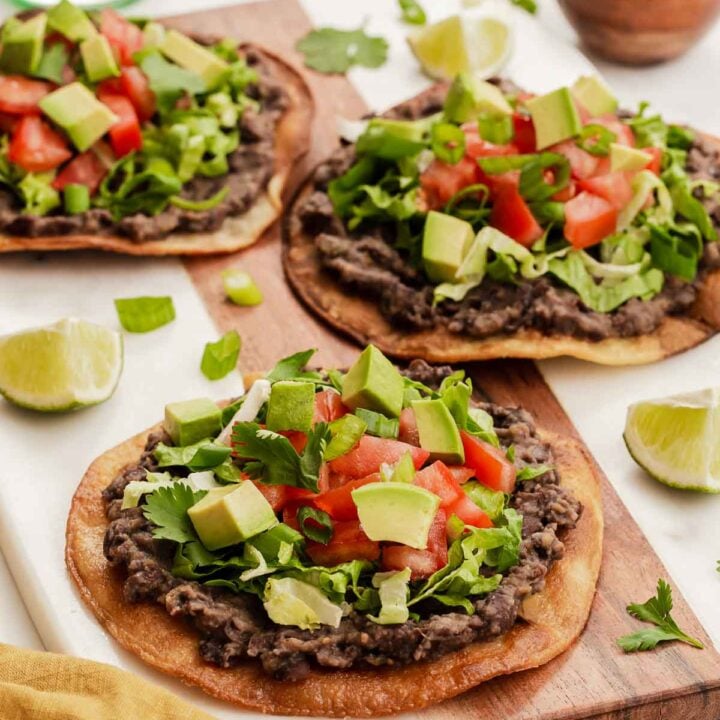 Three black bean tostadas topped with lettuce, avocado and tomatoes on wooden board.