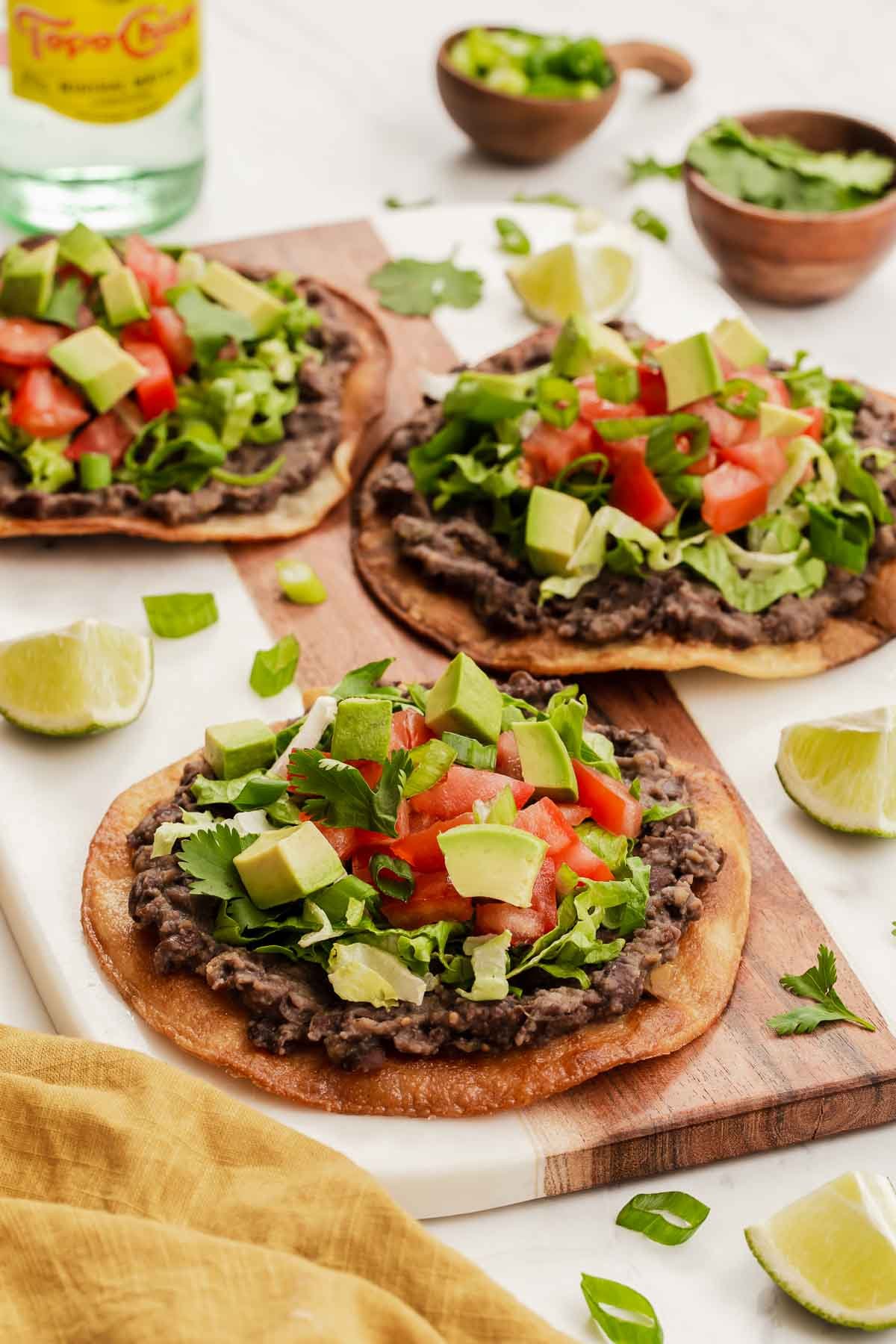 Three black bean tostadas topped with lettuce, avocado and tomatoes on wooden board.