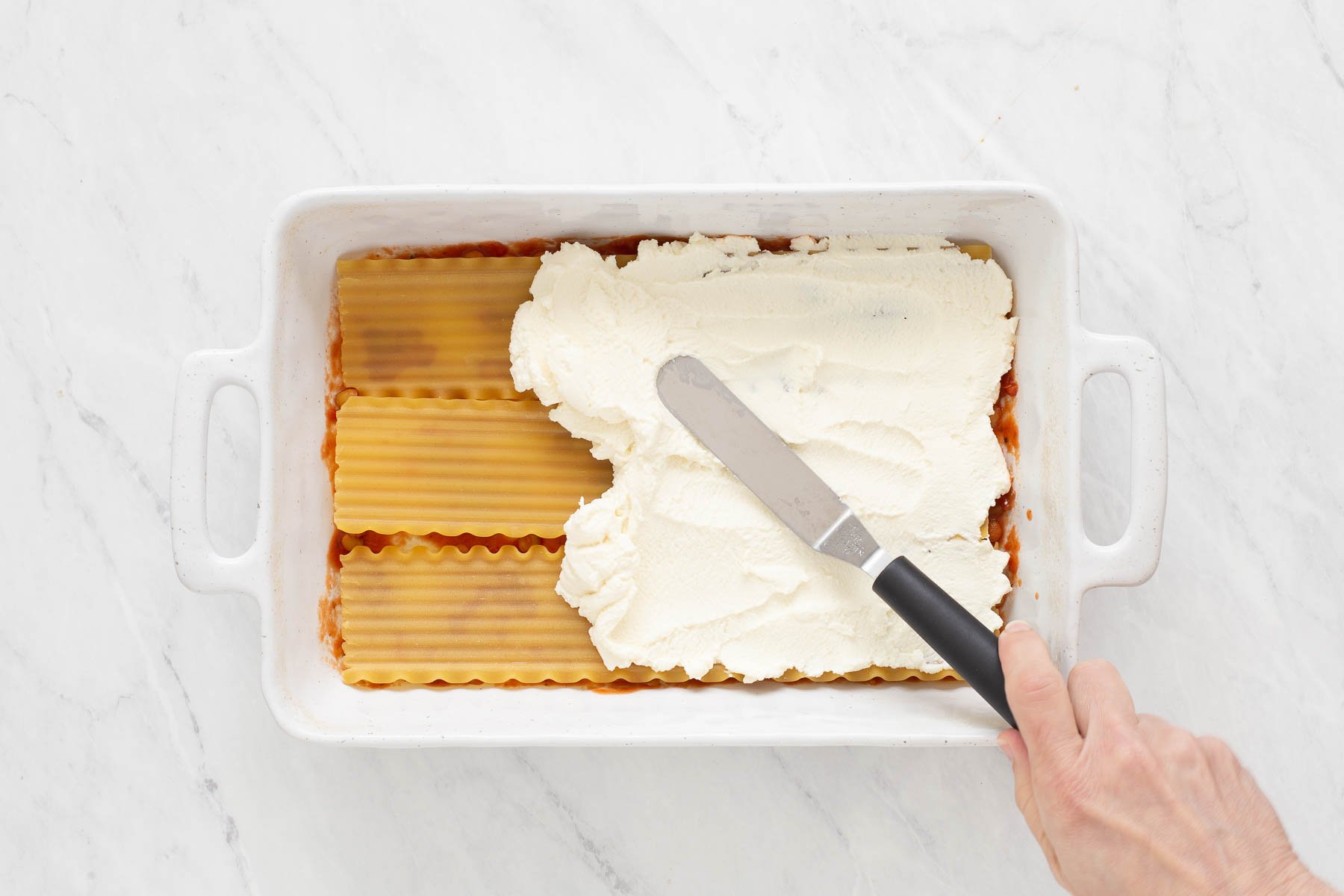 Hand using an offset spatula to spread ricotta in casserole dish.