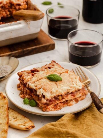 Square slice of lentil lasagna on plate with casserole and red wine behind.