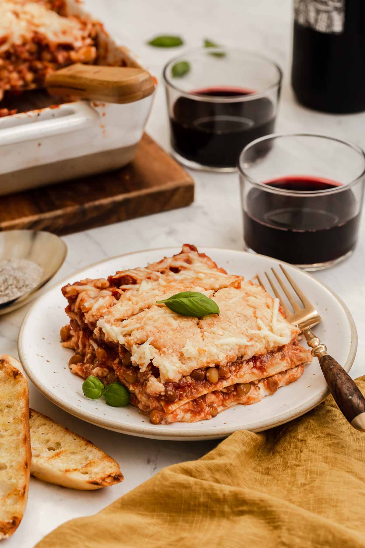 Square slice of lentil lasagna on plate with casserole and red wine behind.