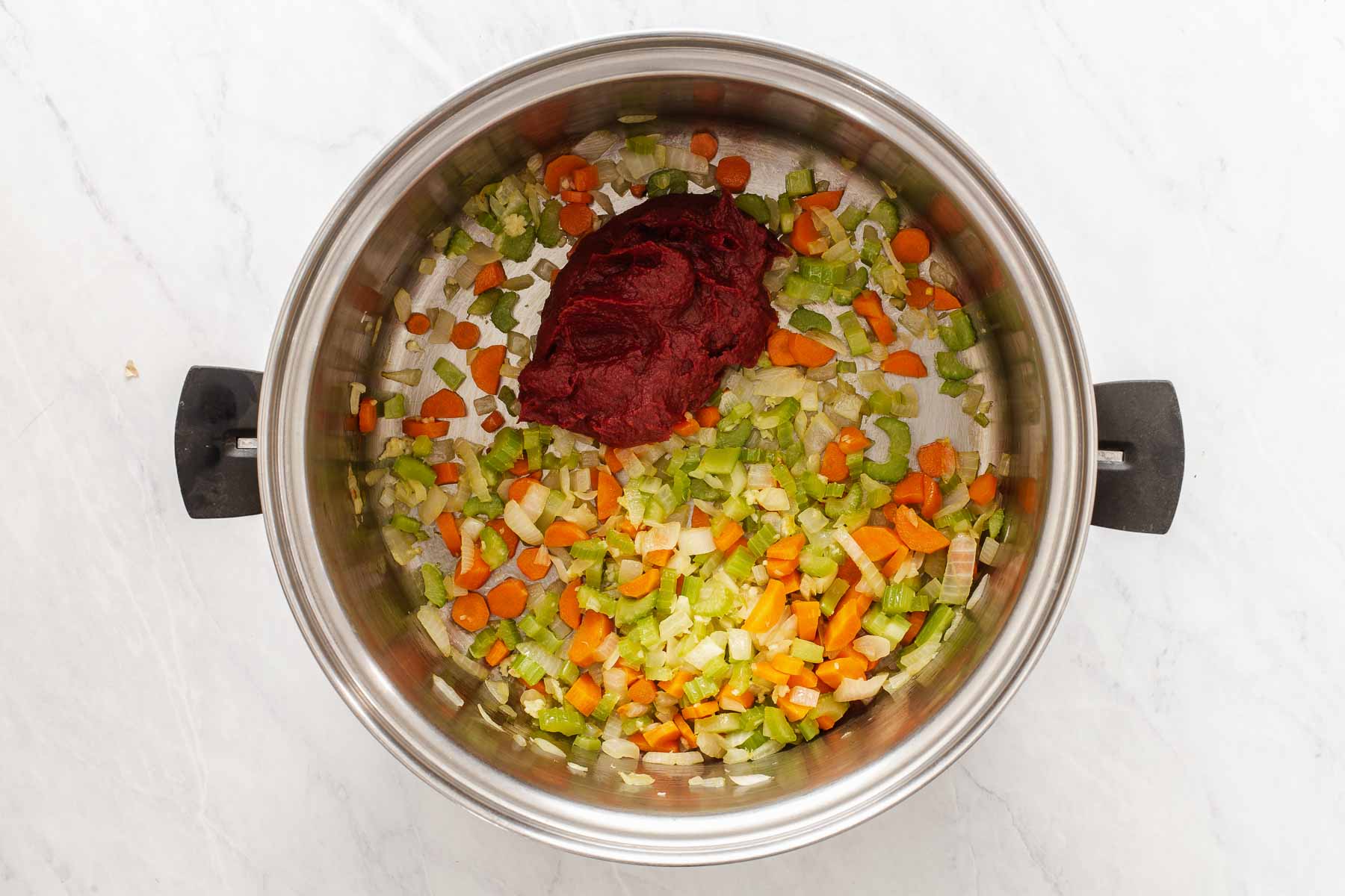 Adding tomato paste to a pot with diced onions, carrots and celery.