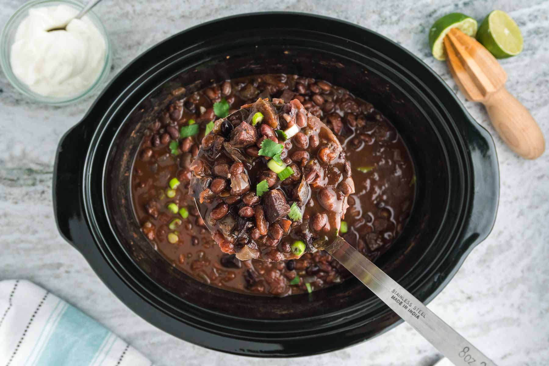 Metal spoon removing a scoop of slow cooker black bean soup from a black crock pot.