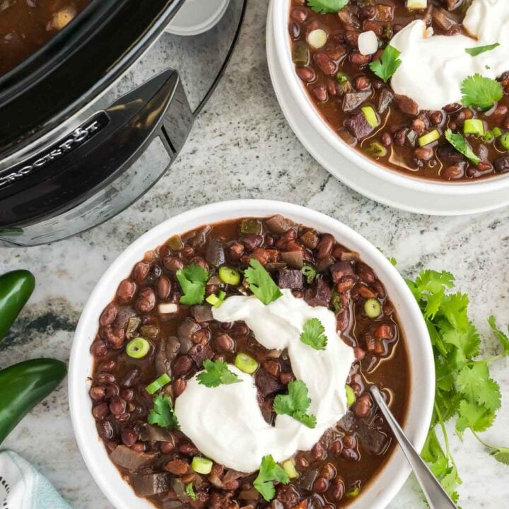 Two white bowls of slow cooker black bean soup next to a crockpot on marble counter.