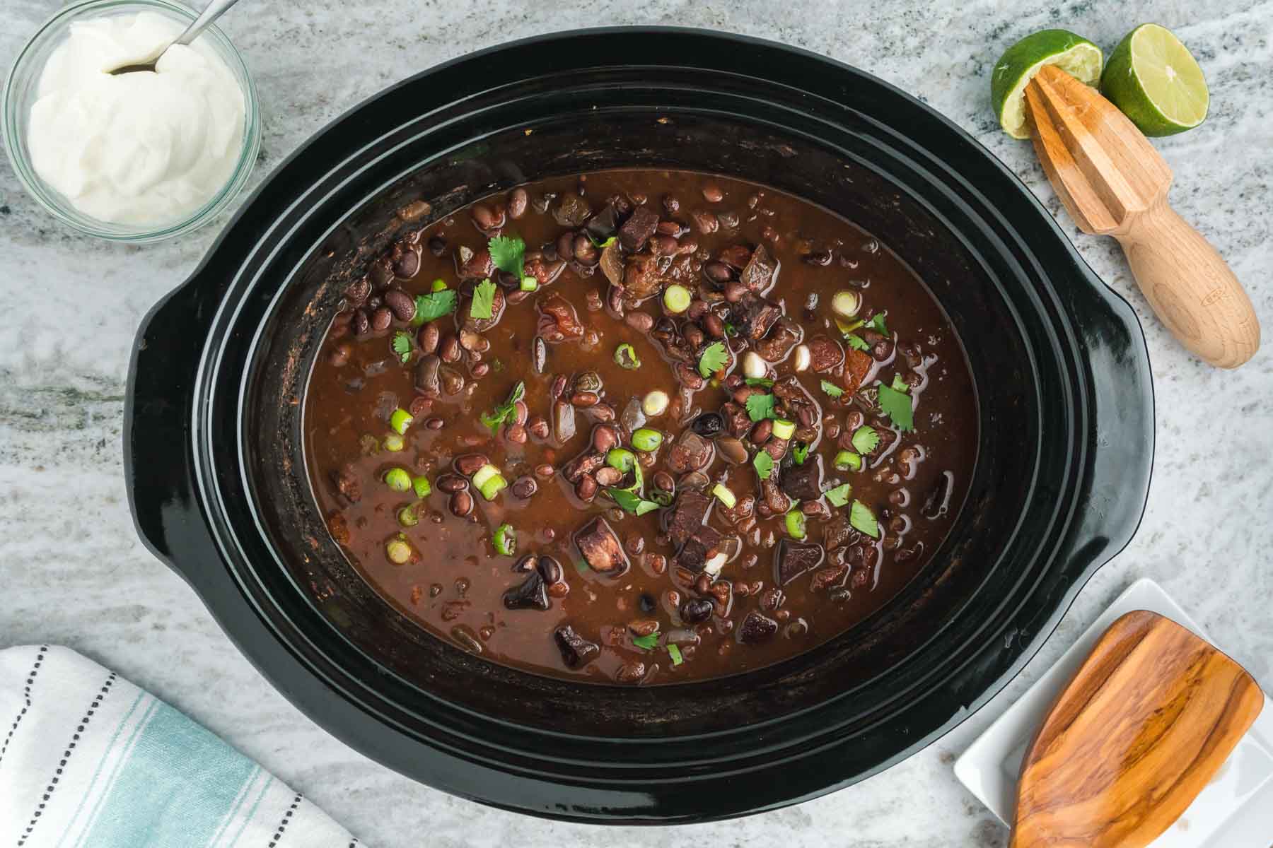 Black slow cooker with black beans in it, garnished with scallions.